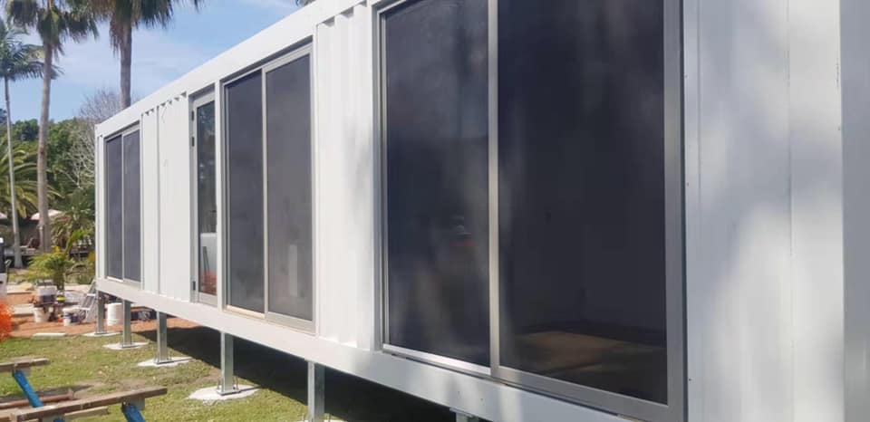White modular building with large, dark windows, elevated on posts, set outdoors with trees.
