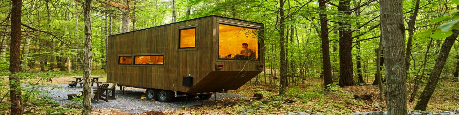 A cabin-like structure on wheels sits in a forest; person inside. Warm light shines from the windows.