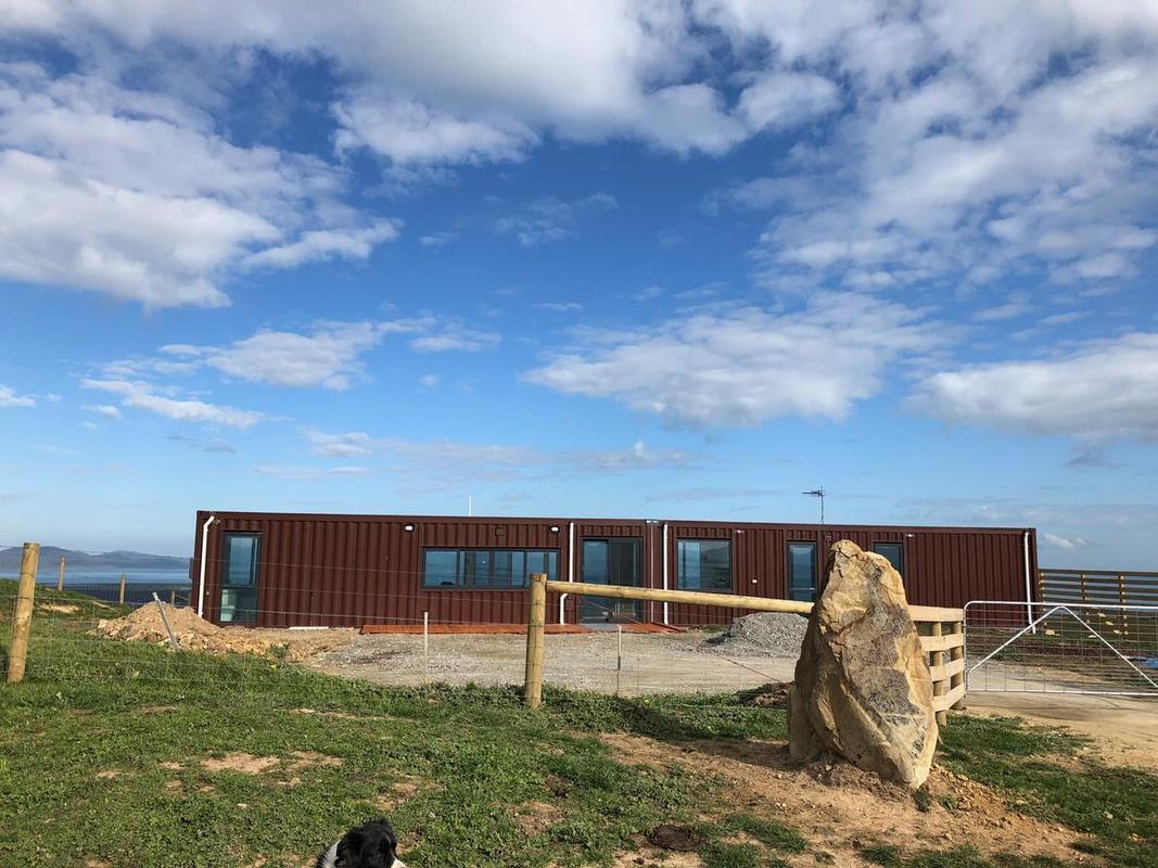 Brown container home on a grassy hill under a blue sky with clouds. A fence and rock are in the foreground.