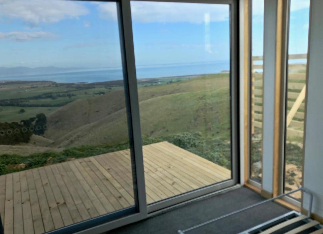 Sliding glass doors open onto a wooden deck overlooking a hilly landscape and ocean under a blue sky.