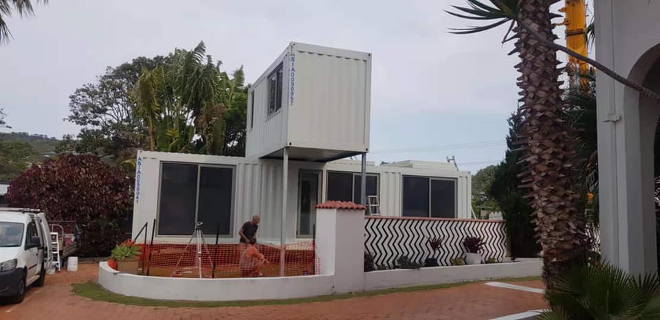 White container home with multiple levels, windows, and a small yard. A palm tree is in the foreground.