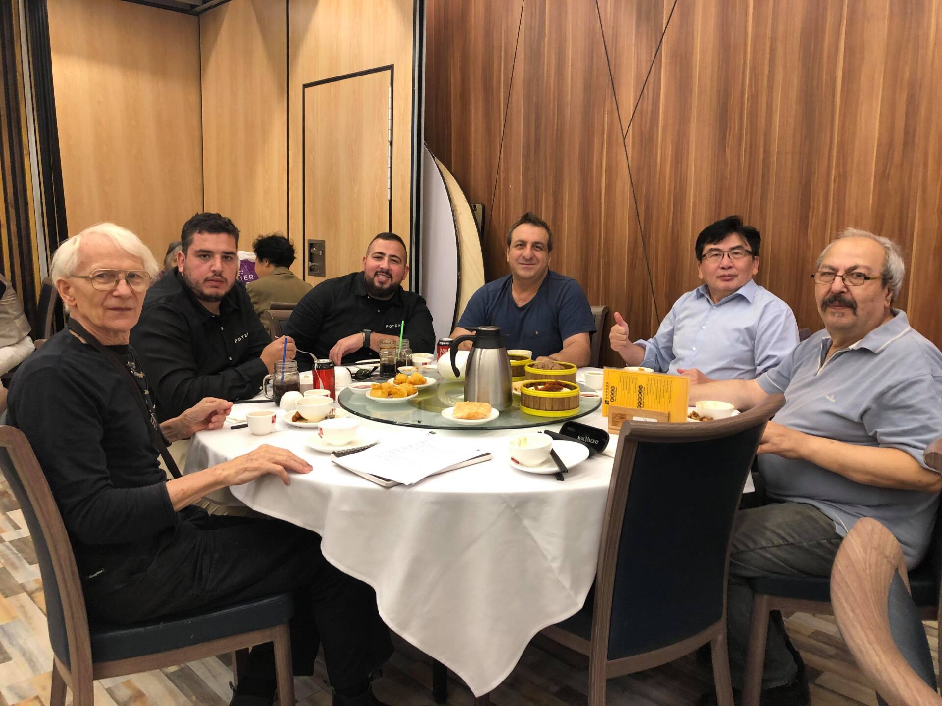A group of men sit around a white tablecloth at a restaurant. They smile and chat over food on the table.