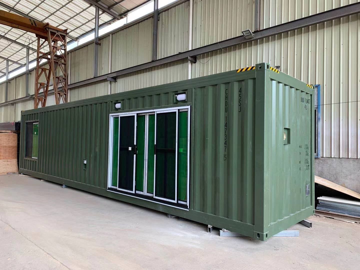 Green shipping container with sliding doors and windows, sitting inside a warehouse.