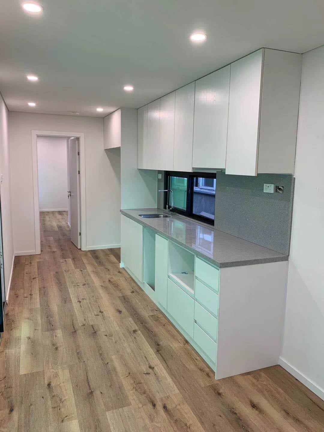 A narrow modern kitchen with white cabinets, light wood floors, and a gray countertop.