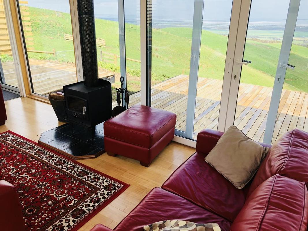 Living room with red leather furniture, wood stove, rug, and large windows overlooking a green landscape.