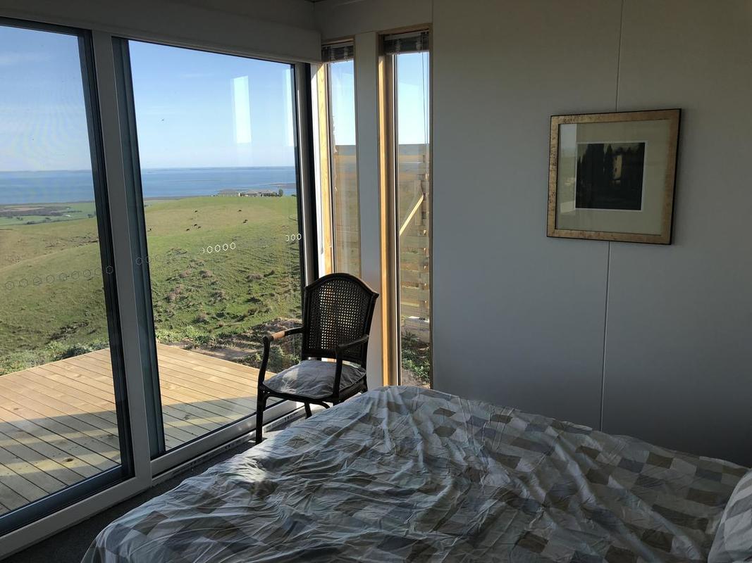 Bedroom with large windows overlooking a green landscape and ocean. Bed, chair, and framed art present.