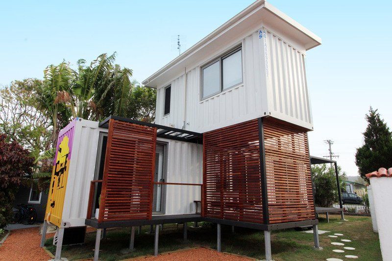 Two-story white container home with wooden accents and a small porch, set in a yard with trees.