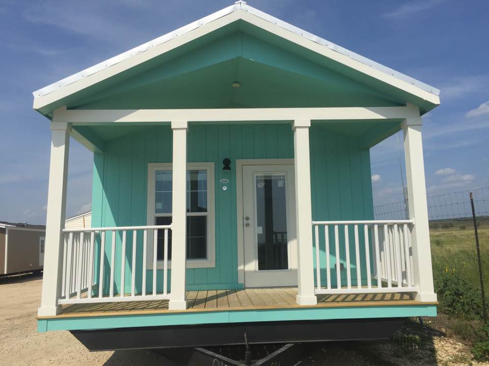 Teal tiny house with white porch, trim, and roof. Wooden deck with open field in the background.