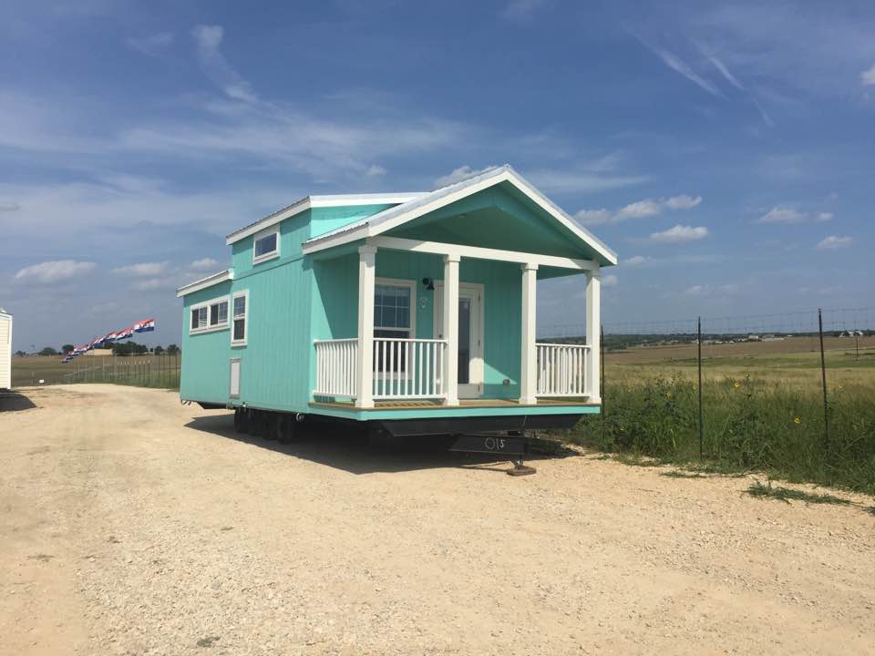 Teal tiny house on wheels with a porch, parked on gravel in a field under a blue sky.