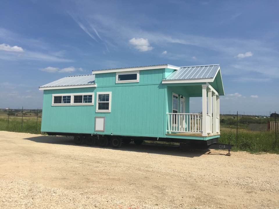 Teal tiny house with white trim, metal roof, and small porch, on gravel under blue sky.