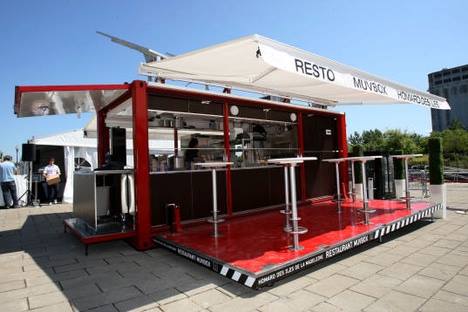 Red mobile restaurant with a white awning, outdoor seating, and stainless steel interior.