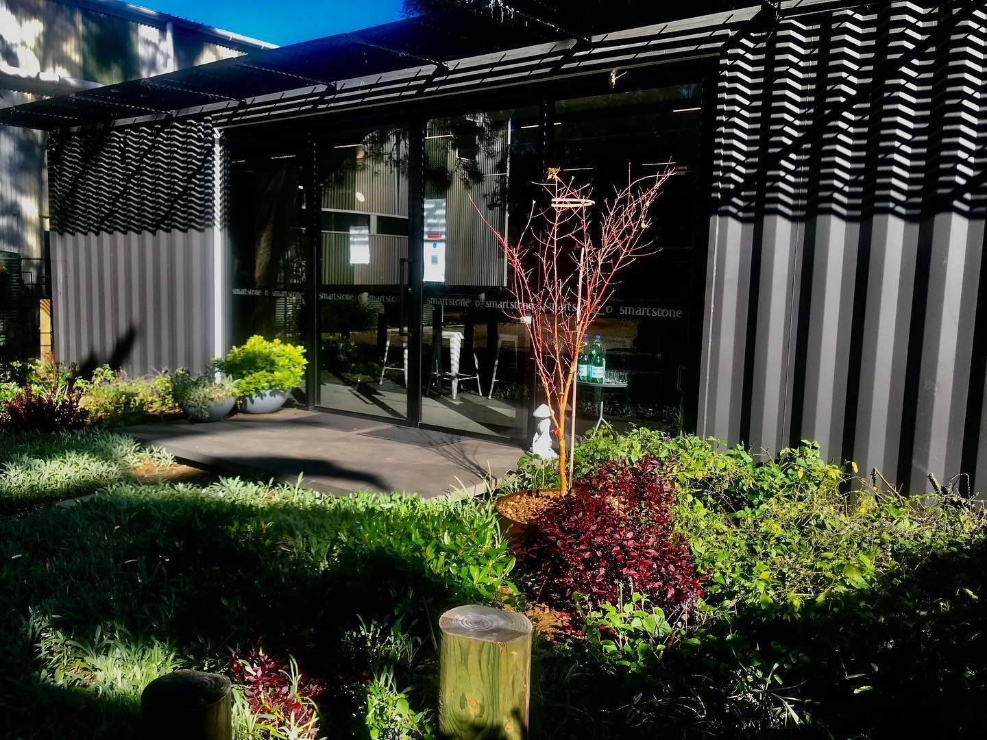 Cafe entrance with glass doors, surrounded by garden beds with green and red foliage.