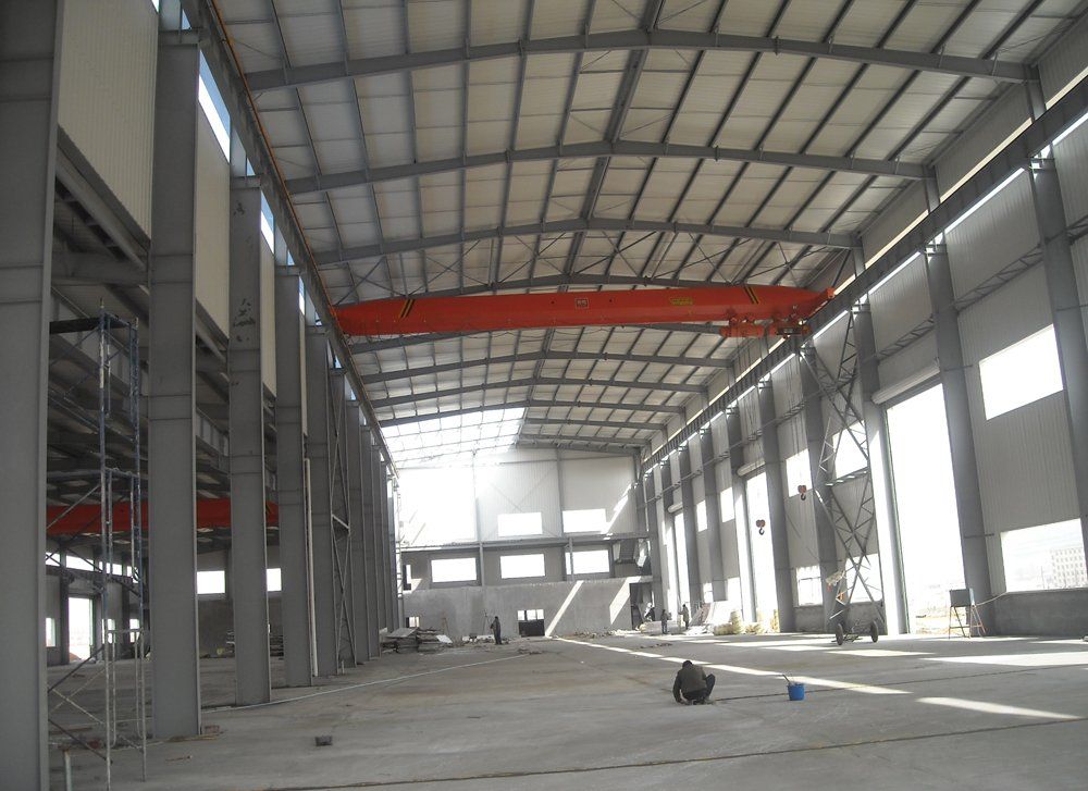 Interior of a large warehouse under construction, steel frame, concrete floor, overhead crane, and natural light.