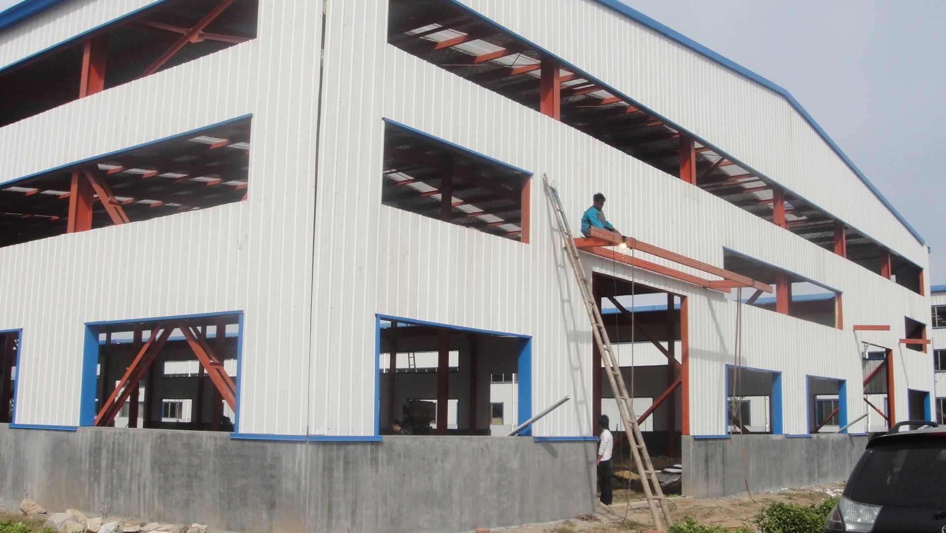 Construction worker on a ladder, applying siding to a large, partially built metal building with blue and white accents.