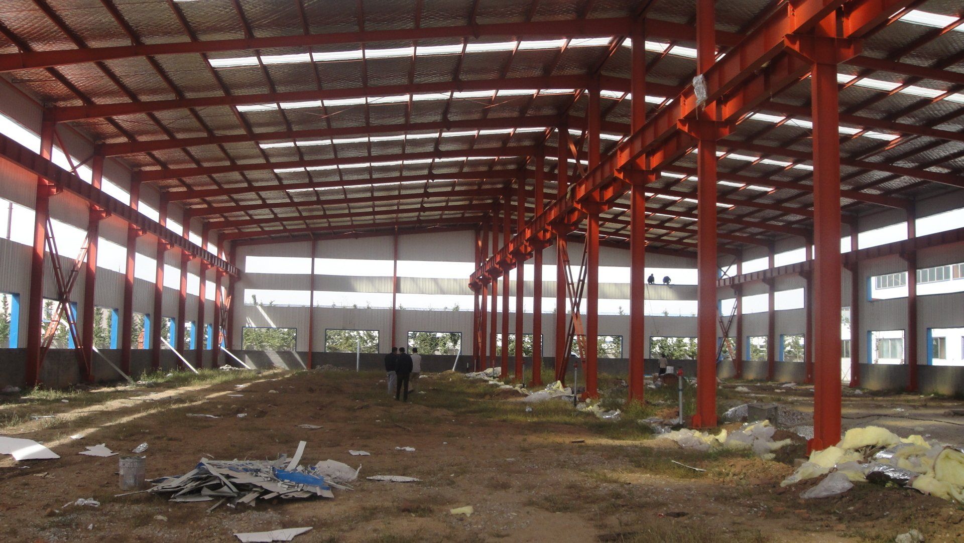 Interior view of an empty industrial warehouse with red metal supports and a person walking towards the back.