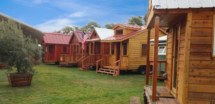 Tiny wooden homes on trailers sit on green grass under a blue sky.