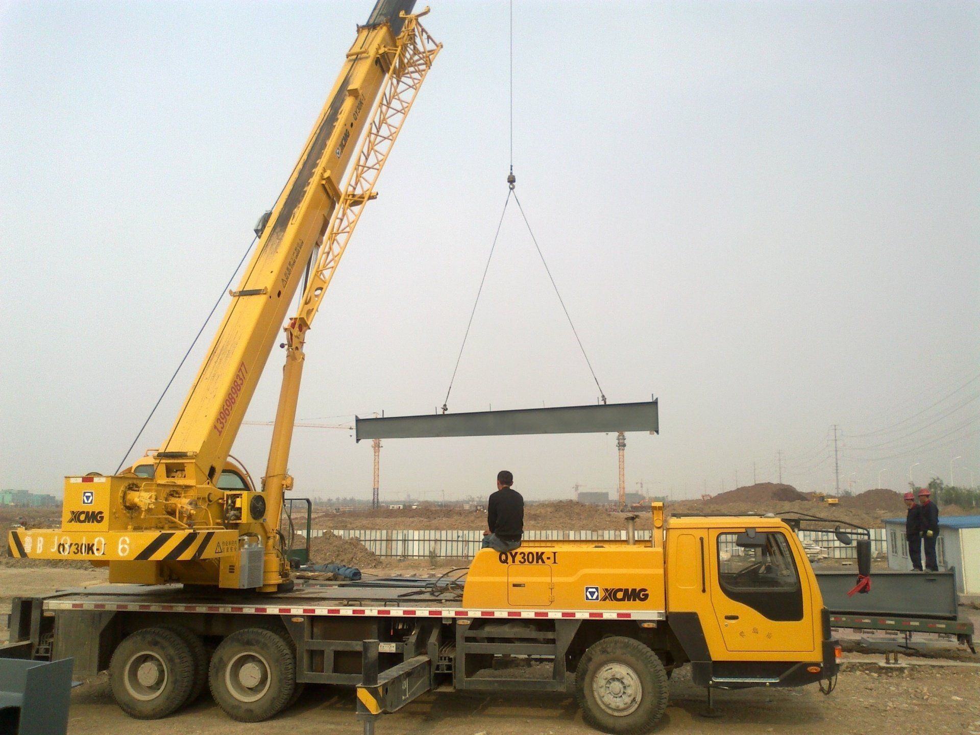 Yellow crane lifting a long, dark beam over a construction site; two workers nearby.