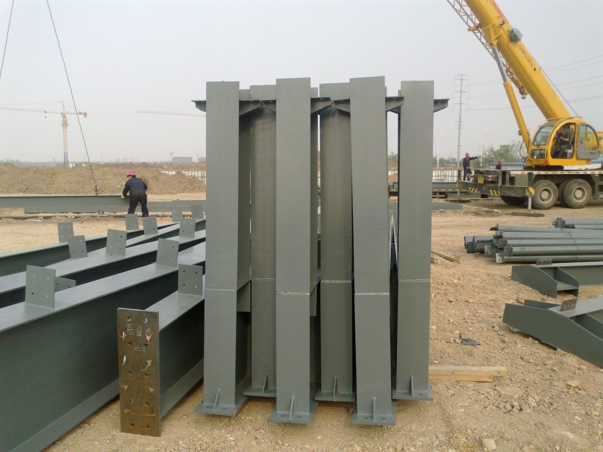 Construction site: gray steel beams and columns, crane in background, worker nearby.