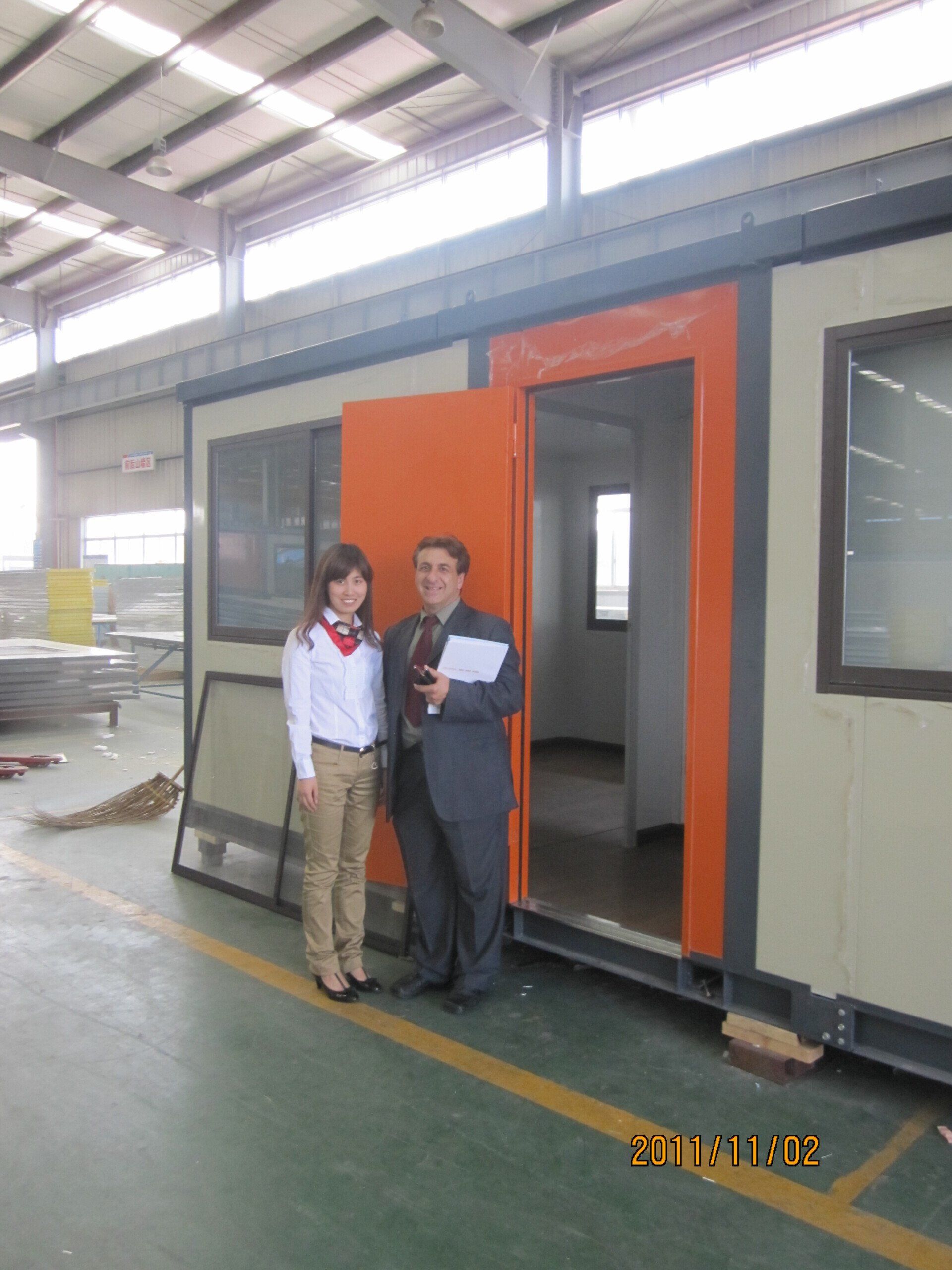 Two people stand in the doorway of a tan modular building with an orange door frame. Inside, another room is visible.