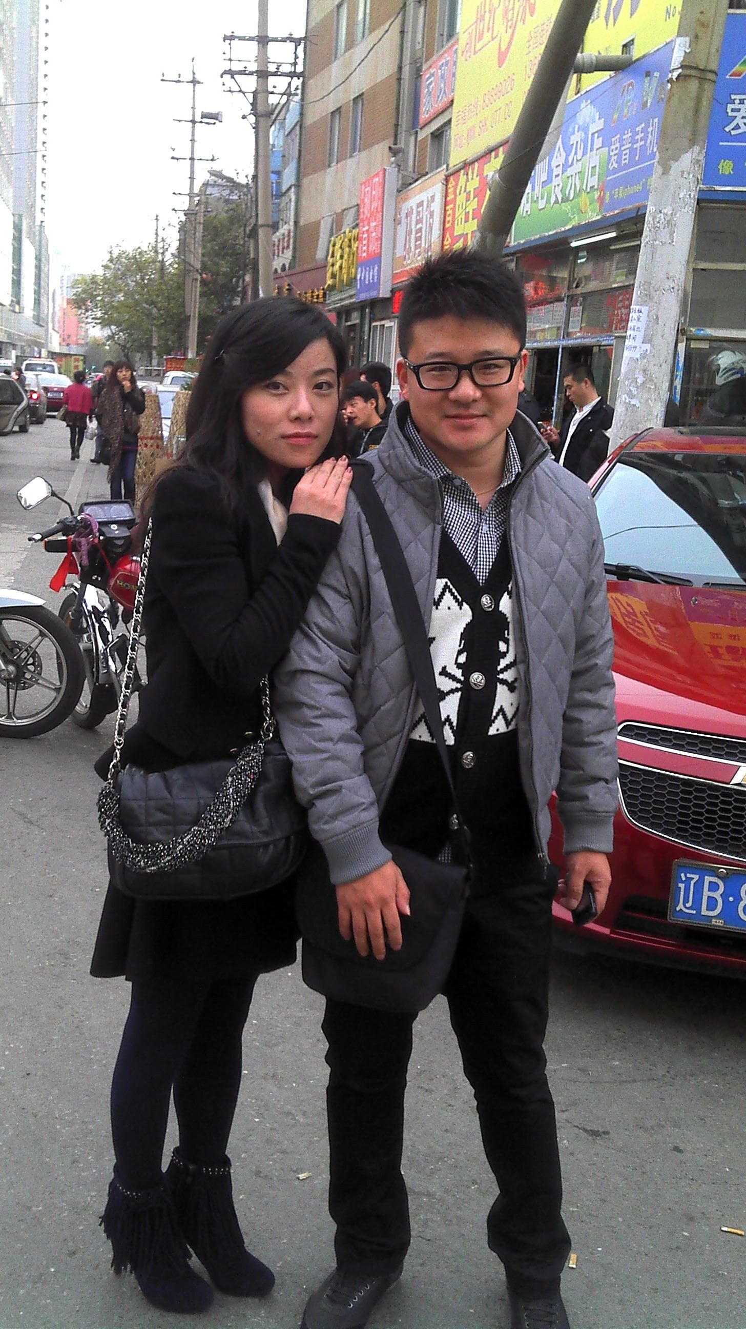 Couple standing on a city street, man in glasses and gray jacket, woman in black coat.