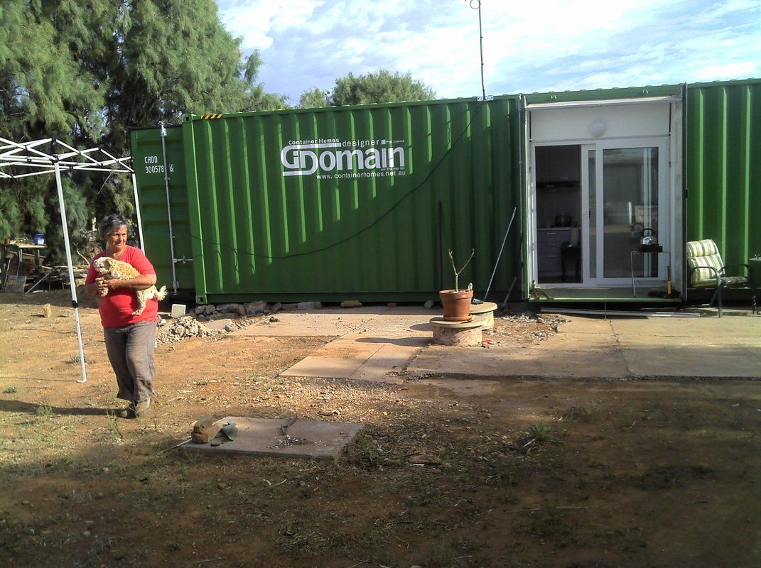 Person holding a small dog stands in front of a green shipping container office with open glass doors.