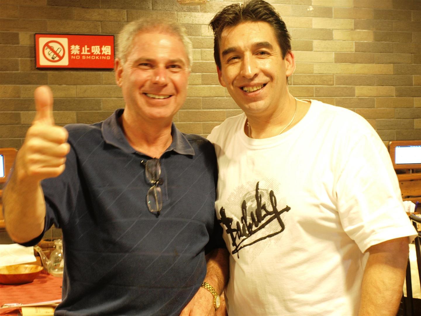 Two men smiling; one gives a thumbs-up. Inside, near a