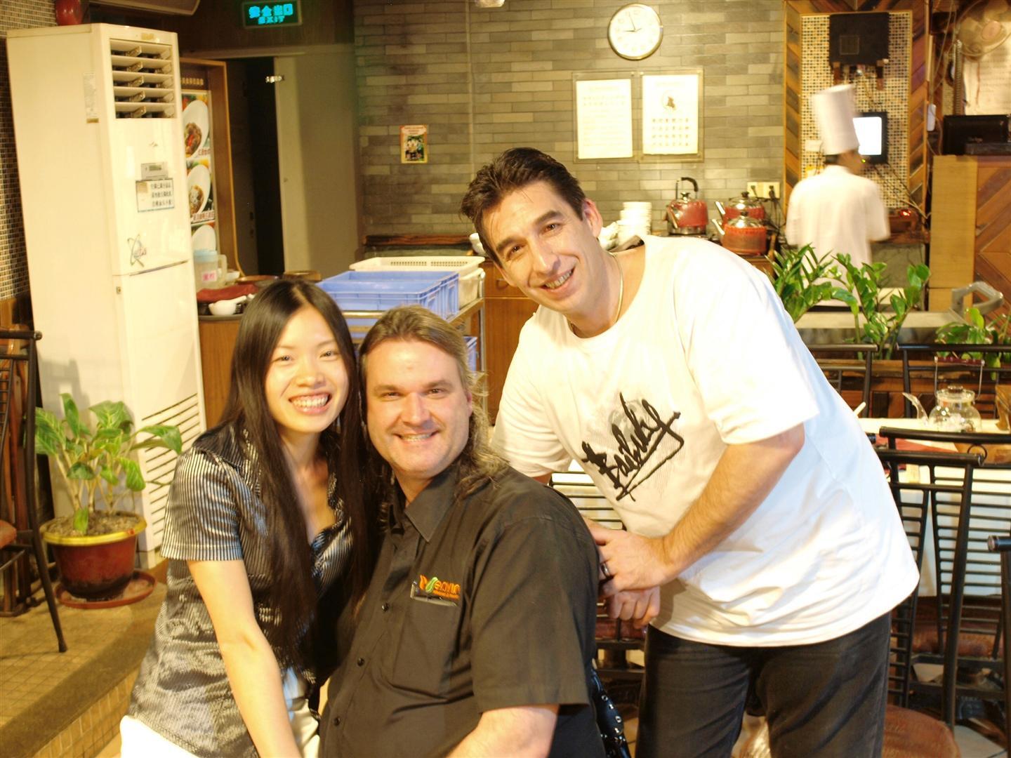 Three people smiling in a restaurant; Asian woman, man, and man wearing a white shirt; indoor setting.