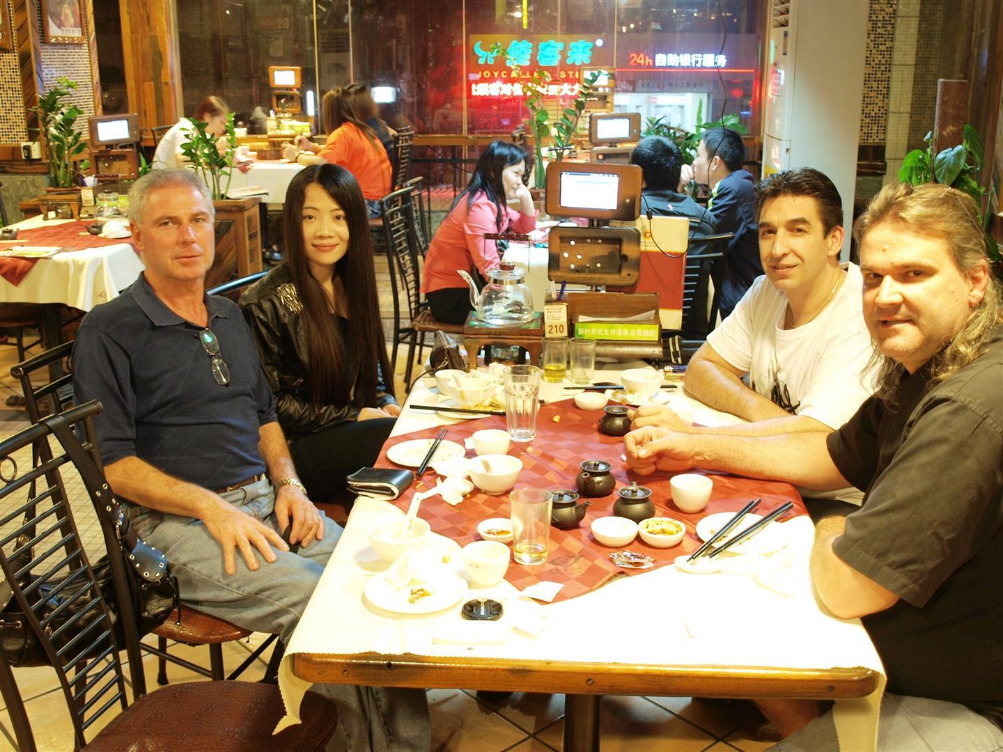 Four people sit around a table in a restaurant. Others dine in the background. Red and white tablecloth.