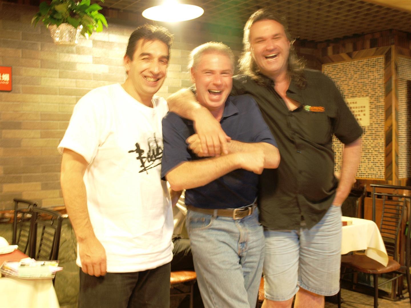 Three smiling men with arms around each other in a restaurant. One wears a white t-shirt, one a blue polo, and one a black shirt.