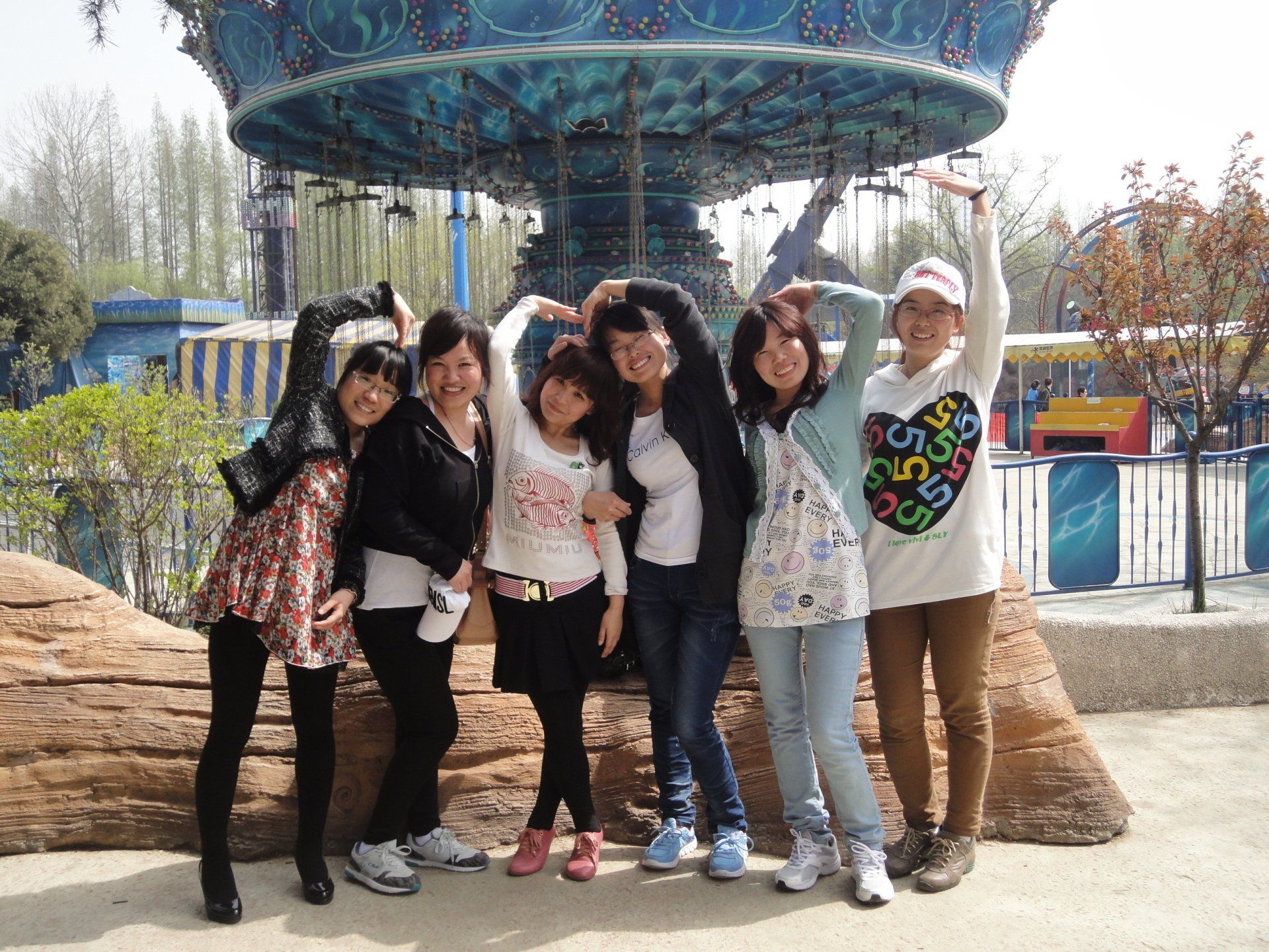 Group of friends pose near a carousel, forming heart shapes with their hands. Amusement park setting.