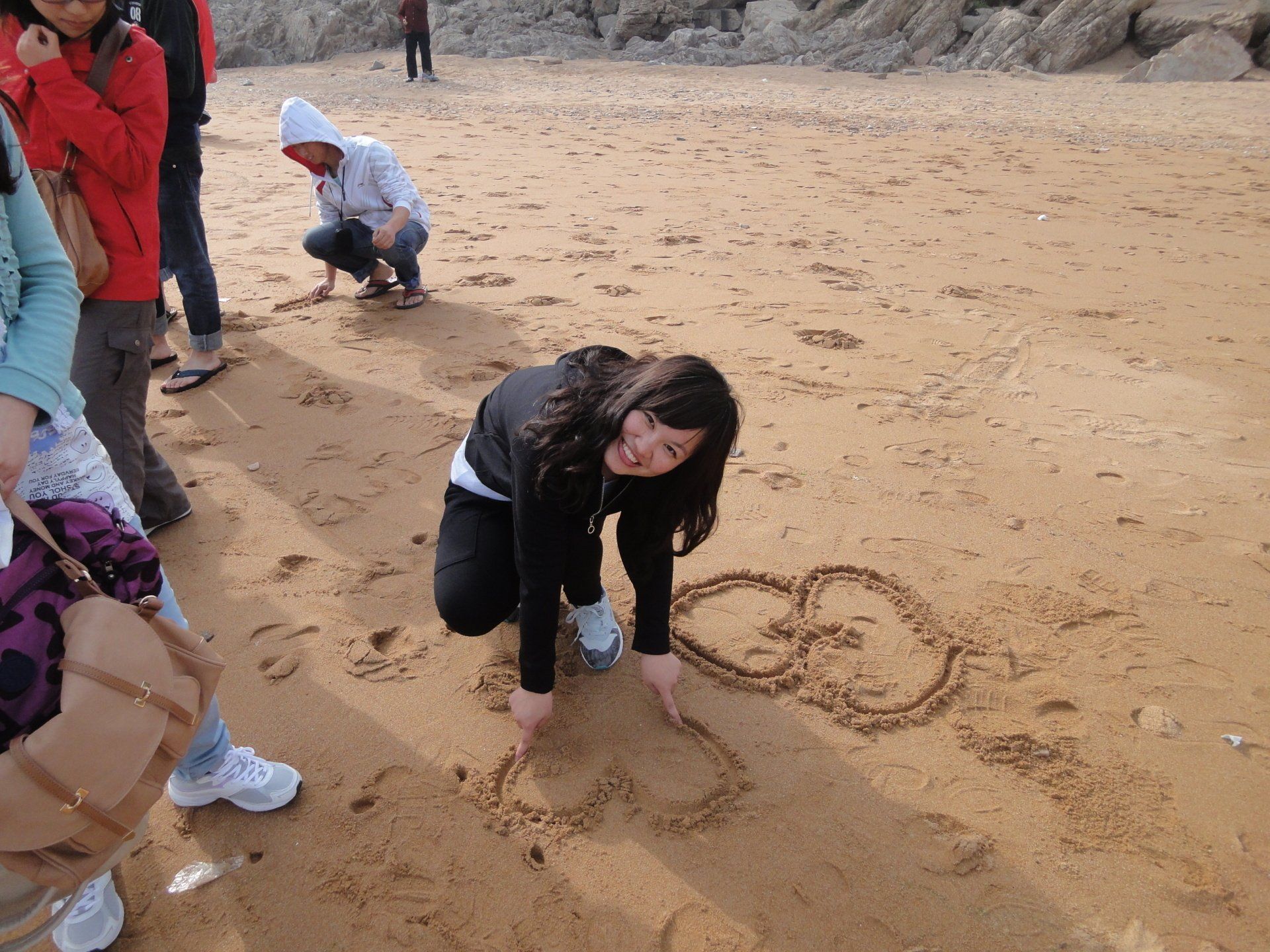 Woman drawing a heart and smiley face in the sand on a beach with other people nearby.