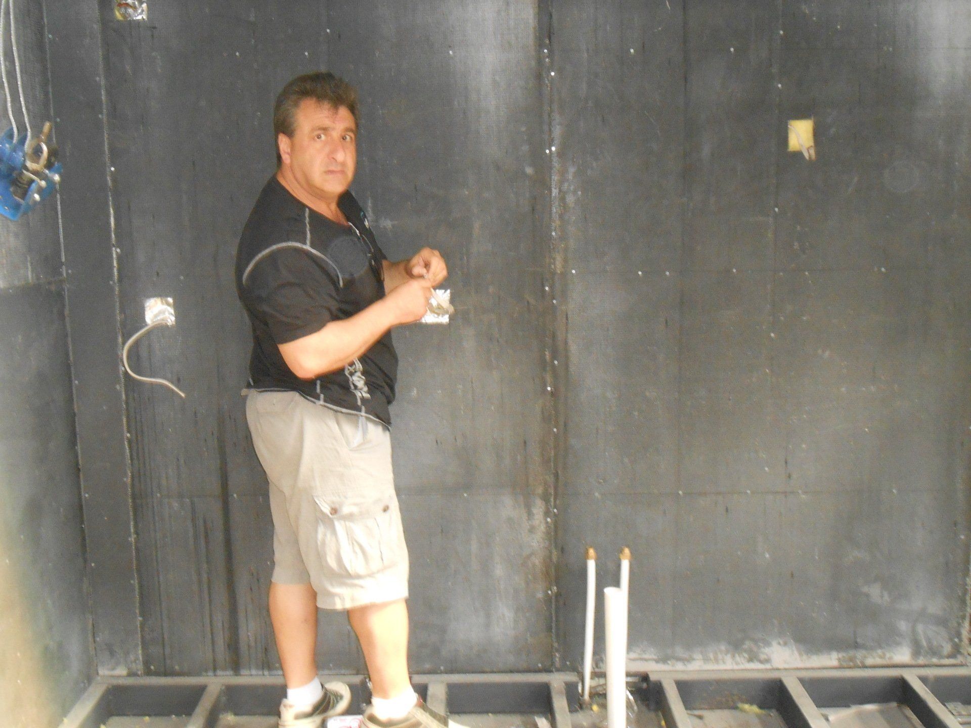 Man in shorts holds electrical component, standing in a room under construction with dark walls and exposed wiring.