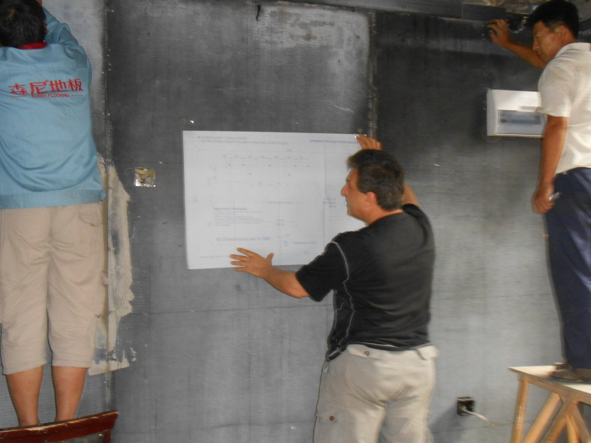 Three people holding a blueprint against a dark wall in what appears to be a construction site.