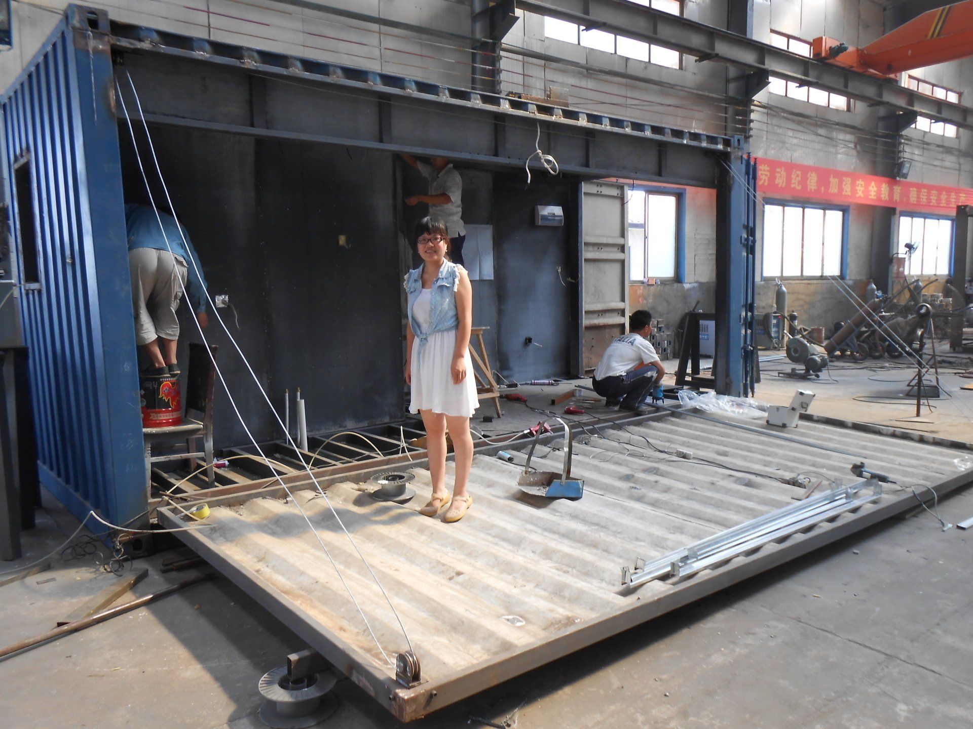 Woman standing inside a partially built container structure in a factory. Men work nearby.