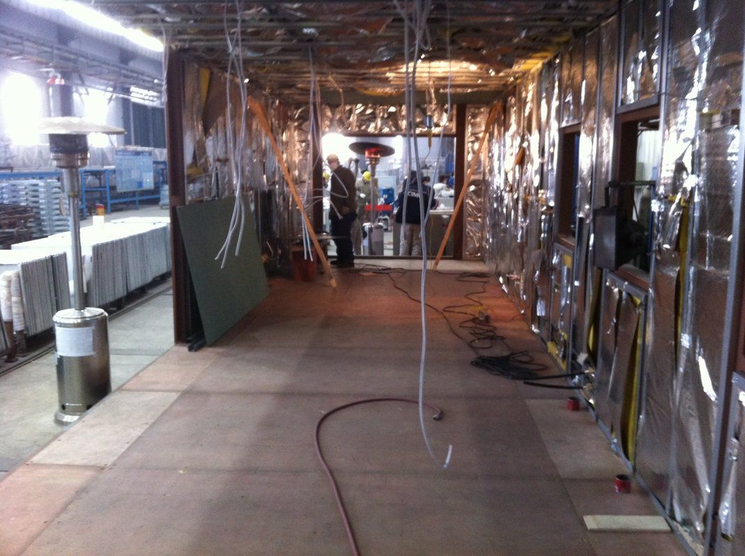 Construction site hallway, walls covered in reflective insulation, workers visible in the distance, propane heater on the left.