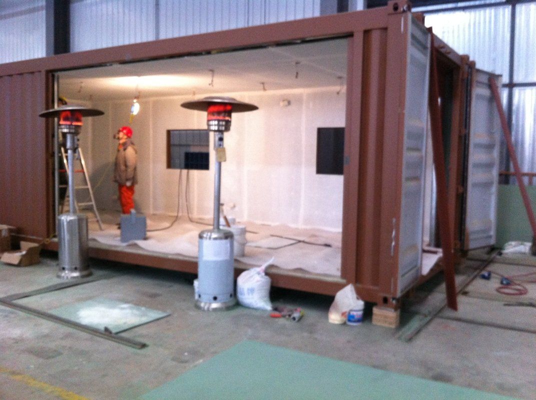 Brown shipping container modified into a room with open sides, gas heaters inside, worker present.