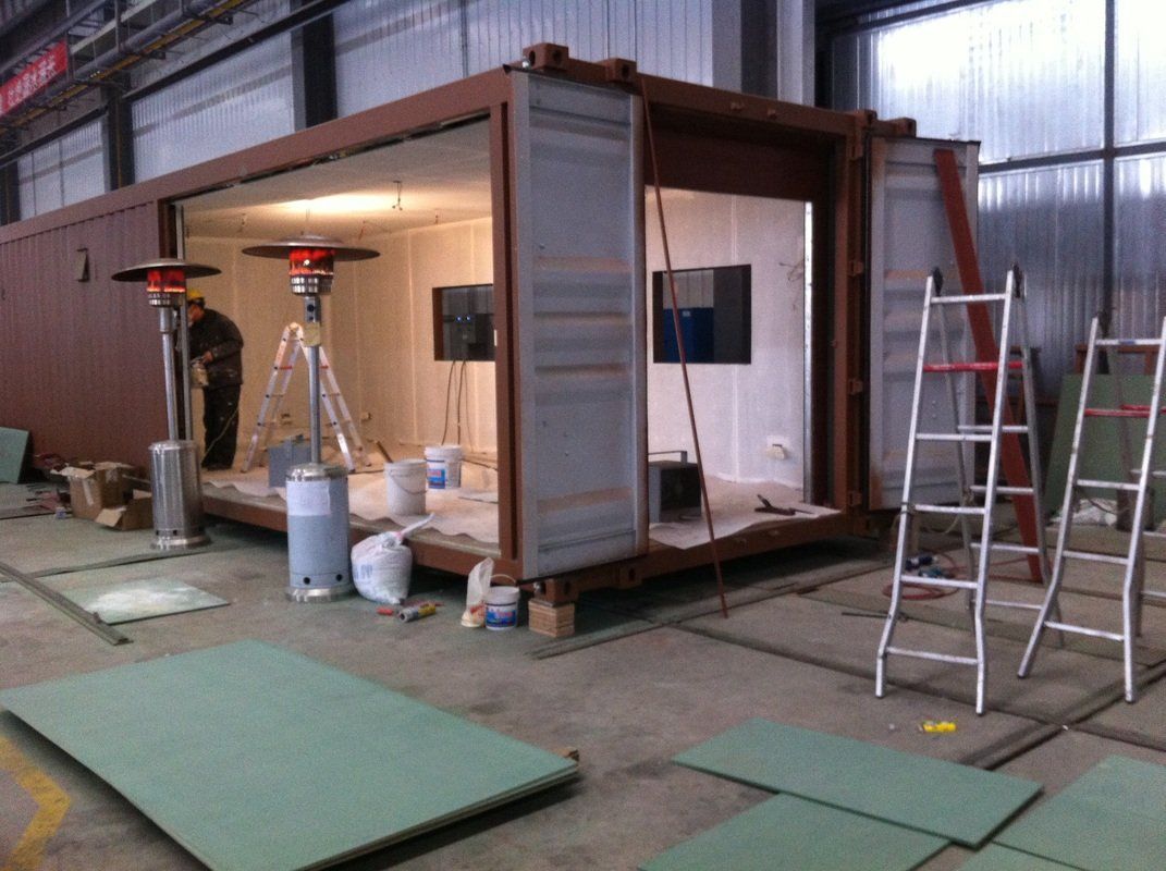 Construction site: interior of a shipping container being renovated, worker, ladders, heaters, paint cans, and drywall.