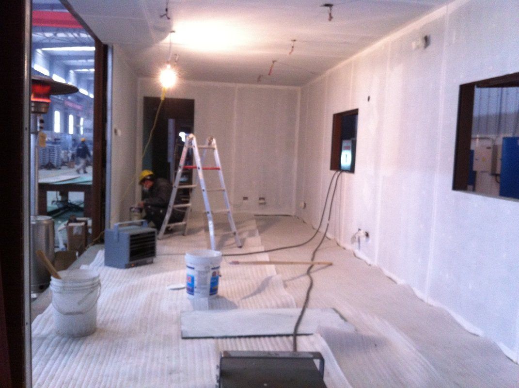 Interior construction scene: Room with white sheeting on walls, protective covering on floor, ladder, and a worker.