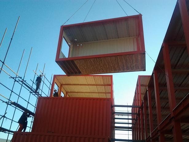 Workers assembling orange shipping containers with scaffolding under a blue sky.
