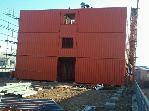 Orange shipping containers stacked to form a three-story building under construction, with scaffolding and cut-out openings.