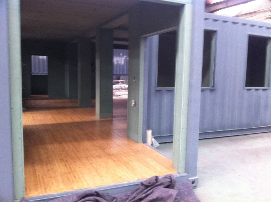 Interior of a repurposed shipping container with a wooden floor and window frames. Walls are gray-green.