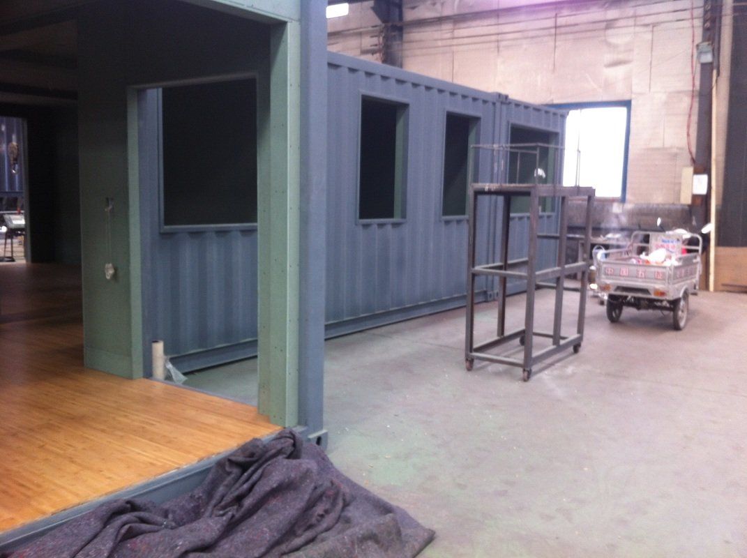 Blue shipping containers with windows, connected, under construction in a warehouse.