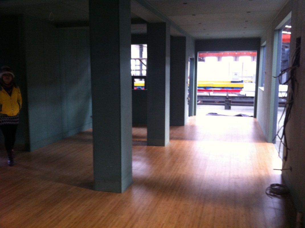 Empty interior with wood floor and teal pillars. Open doorway leads to street with a yellow vehicle.