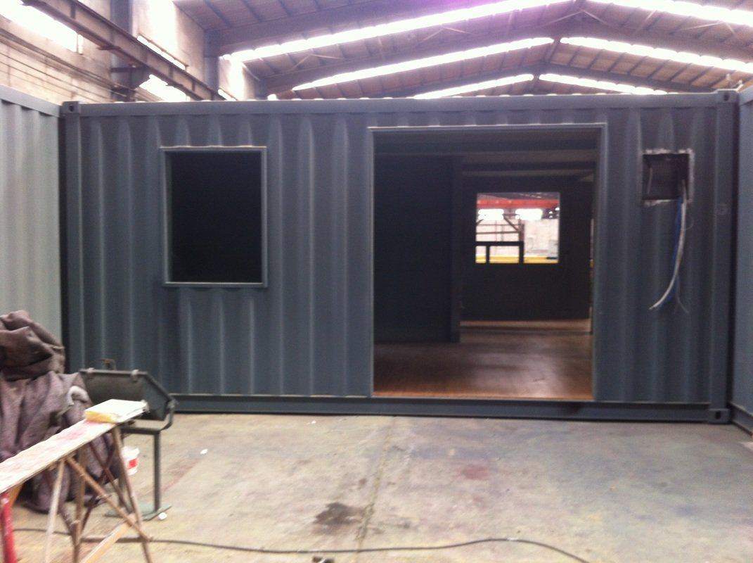 Shipping container modified with windows and doorways, interior view. Dark gray exterior, wooden floor, industrial setting.