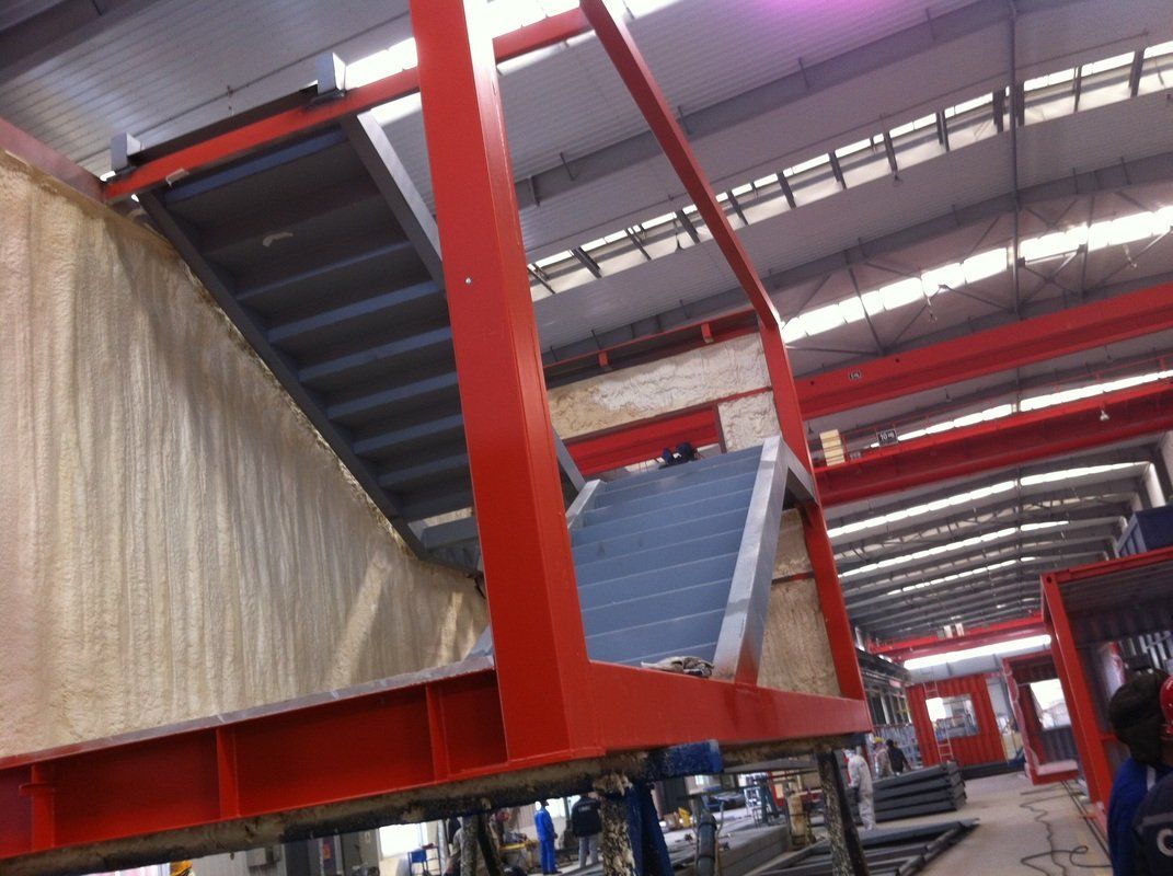 Red metal framework supporting a staircase inside a warehouse, with corrugated metal steps and beige wall.