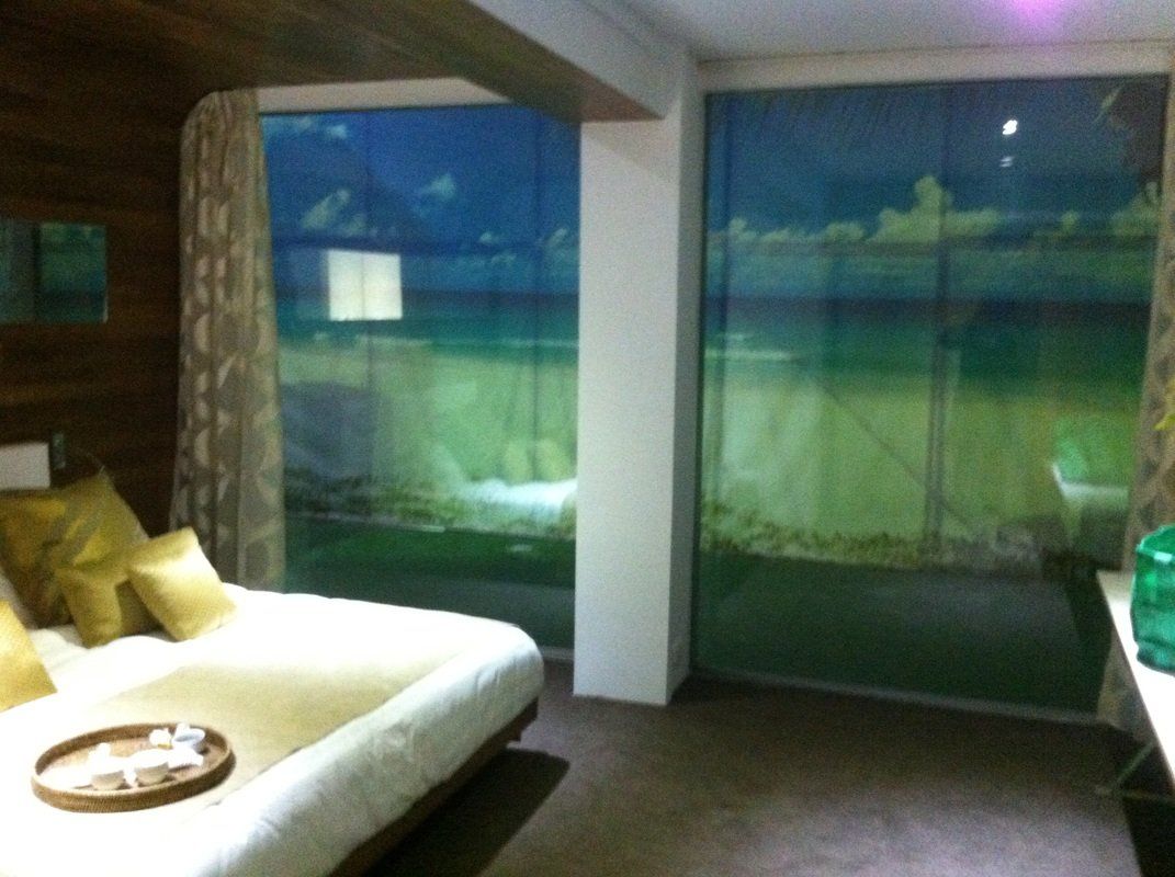Bedroom with bed and large windows displaying beach scene. Green, blue, and brown hues.