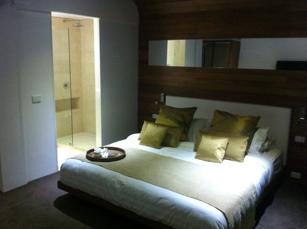 Bedroom with a large bed, gold pillows, wooden accent wall, and a glass-enclosed shower visible.