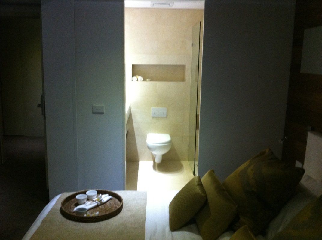 Bedroom doorway leads to bathroom with toilet and built-in shelf. Beige and white tones.