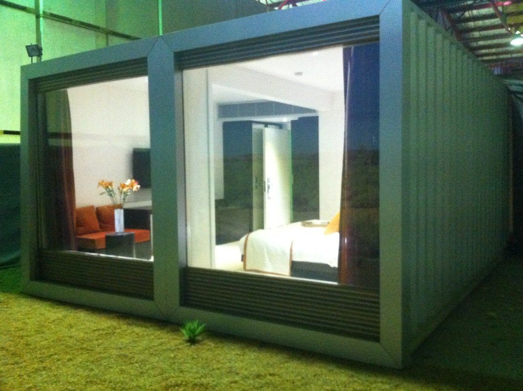 Modern container home with large glass windows, revealing a furnished interior; silver exterior on green grass.