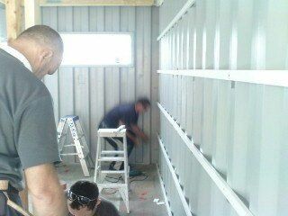 Men installing vertical siding on a wall with a window. One is on a ladder, others work below.